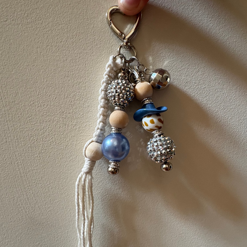 A handmade beaded keychain charm held by a hand against a beige textured background. The keychain features a silver metal heart clasp, a white braided cord, a white tassel, wooden beads, a blue bead, a blue hat charm, and silver rhinestone-covered beads.