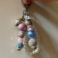 A handmade beaded keychain charm held by a hand, featuring various colorful beads, a pearl strand, and a small daisy flower on a beige background.