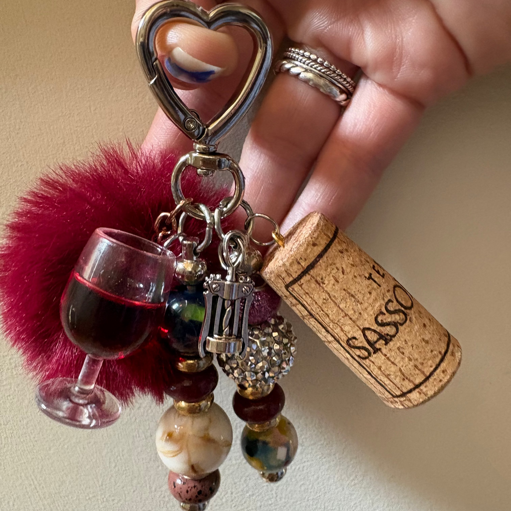 A close-up of a hand holding a keychain charm set. The keychain features a silver heart-shaped clasp, a magenta fuzzy pom-pom, a miniature wine glass charm filled with red liquid, a cork charm printed with 'SASSO', and several dangling beads in maroon, cream, and patterned glass.