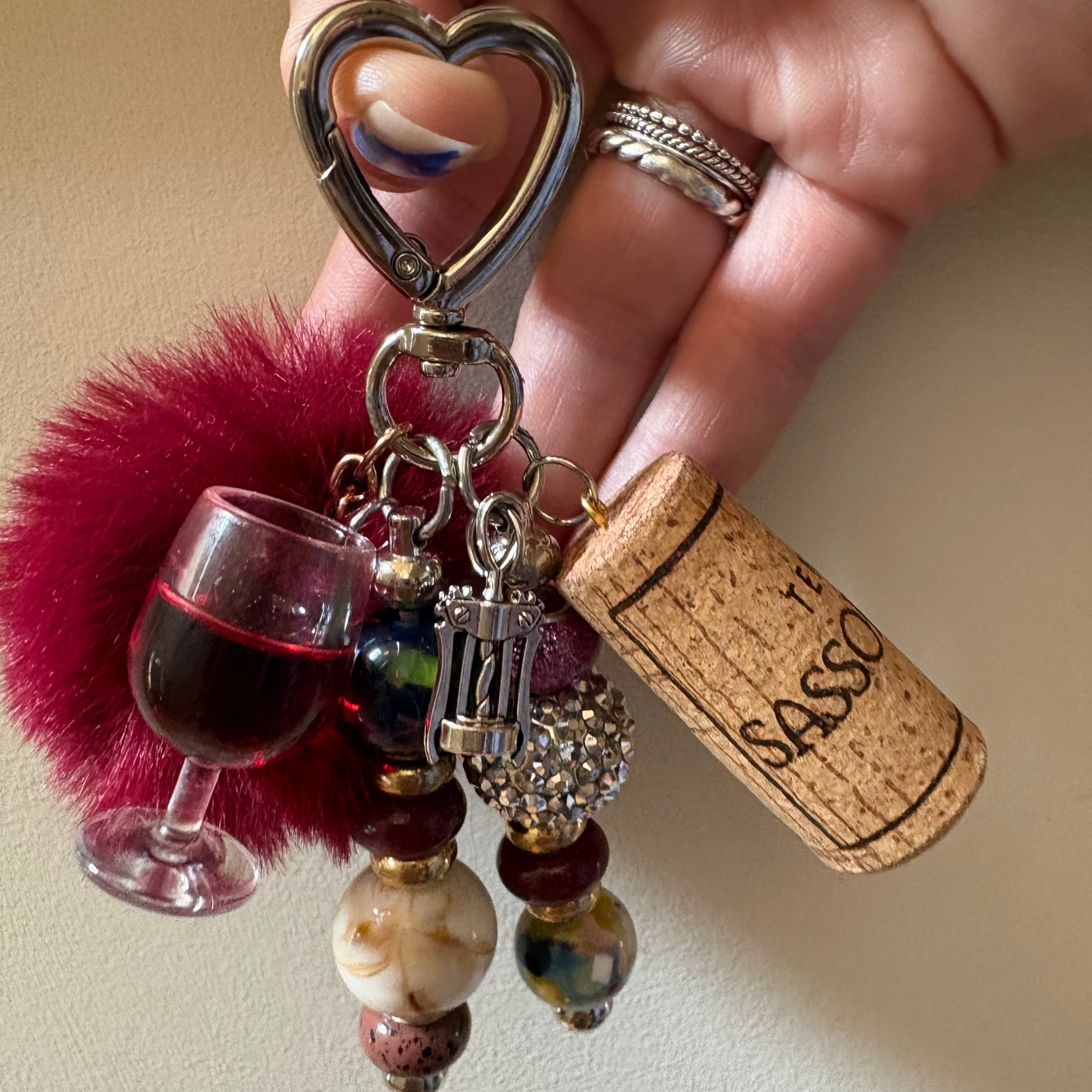 A close-up of a hand holding a keychain charm set. The keychain features a silver heart-shaped clasp, a magenta fuzzy pom-pom, a miniature wine glass charm filled with red liquid, a cork charm printed with 'SASSO', and several dangling beads in maroon, cream, and patterned glass.