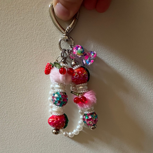 A close-up of a handmade keychain charm held by a hand, featuring colorful beadwork, faux pearls, pink pom-poms, and strawberry embellishments on a silver key ring, set against a plain beige wall.